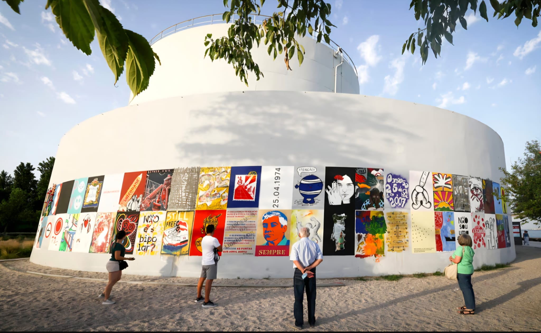 Pessoas observam um mural de cartazes numa fachada branca curva, ao ar livre.